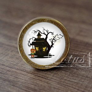 Pode incluir: Um botão de armário redondo de cor latão com um inserto de cerâmica branca que apresenta uma ilustração de Halloween de uma casa assombrada com uma abóbora esculpida na frente.