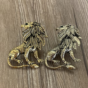 May include: Two gold-toned lion brooches with green gem accents. The lions are detailed with intricate leaf patterns and have a textured finish.