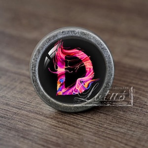May include: A silver cabinet knob with a round, black base and a glass top featuring a colourful abstract design of the letter "D".