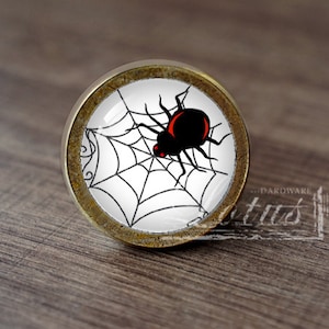 Pode incluir: Um botão de armário redondo de cor latão com um inserto de vidro que apresenta uma aranha preta com olhos vermelhos numa teia branca.