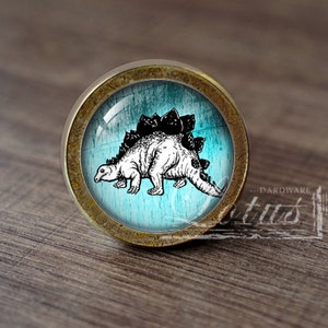 May include: A round, brass-colored cabinet knob with a glass insert featuring a black and white illustration of a stegosaurus on a blue background.