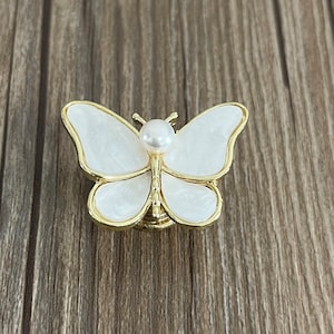 May include: A white butterfly-shaped drawer pull with a gold trim and a pearl accent in the center.