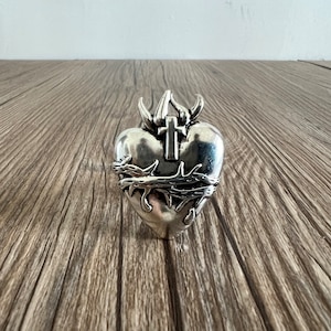 May include: A silver-colored heart-shaped pendant with a cross and a crown of thorns. The pendant has a detailed design and is likely made of metal. The heart shape is prominent, and the overall aesthetic is religious.
