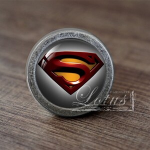 Könnte beinhalten: Ein silberfarbener Schrank-Knopf mit einer runden, gläsernen Oberfläche, die das rote und gelbe Superman-Logo zeigt.