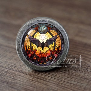 May include: A round, silver-toned cabinet knob with a stained glass design featuring a bat flying in front of a full moon. The stained glass design has a yellow, orange, and red colour scheme.