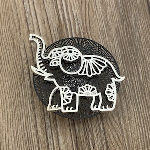 以下が含まれることがあります： 黒いマンダラのような背景に、繊細な透かし模様が施された銀色の象のチャーム