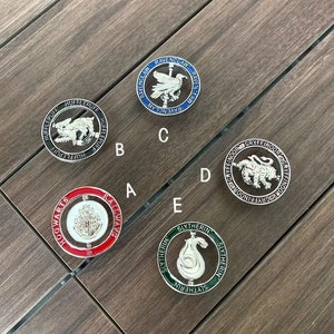 May include: Five round metal badges with different coloured backgrounds and silver accents. Each badge features a different Hogwarts house crest with the house name written around the crest. The crests are: Gryffindor, Hufflepuff, Ravenclaw, Slytherin, and Hogwarts.