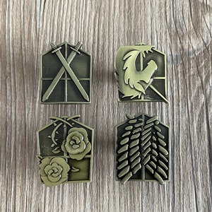 Peut inclure: Quatre épingles en métal de couleur bronze avec des motifs différents. La première épingle représente deux épées croisées, la deuxième épingle représente une créature ailée, la troisième épingle représente une rose et des vignes, et la quatrième épingle représente une série de plumes.