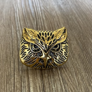 May include: A gold-coloured owl ring with black eyes and detailed feathers. The ring is made of metal and has a textured surface.