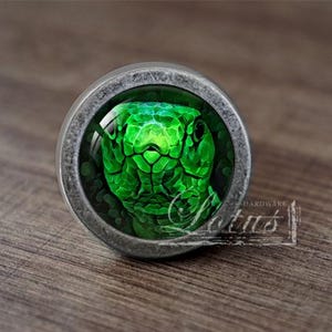 May include: A silver-toned cabinet knob featuring a green snake head design. The snake's eyes are black and the background is a dark green.