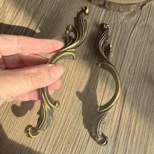 以下が含まれることがあります： 装飾的で曲線的なデザインの、アンティークスタイルのブロンズ製キャビネットハンドル2つ。ハンドルは、金色のハイライトが入った濃いブロンズ仕上げで、装飾的な花のモチーフが特徴です。家具やキャビネットに適しています。