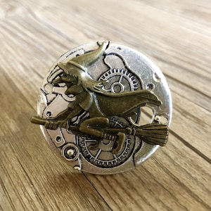 Op de afbeelding: Een zilverkleurige metalen pin met een gedetailleerd ontwerp van een heks die op een bezem rijdt. De heks draagt een punthoed en een lange, vloeiende jurk. De pin is een grillig en decoratief stuk.