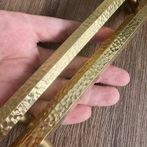 May include: A pair of gold hammered metal cabinet pulls with a rectangular shape. The pulls have a textured surface and are designed to be used as hardware for cabinets or drawers.