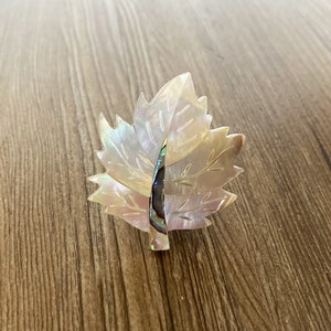 May include: A white and iridescent mother-of-pearl leaf brooch. The leaf is made of two overlapping pieces of shell, with a detailed vein pattern. The brooch is a delicate and elegant piece of jewellery.