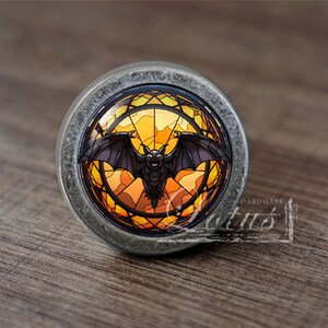 May include: A round, silver-toned cabinet knob with a stained glass design featuring a black bat in a yellow and orange circular frame.