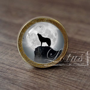 Può includere: Un pomello per armadio rotondo in ottone con un inserto in vetro che presenta una silhouette di un lupo che ulula alla luna piena. La luna è bianca e lo sfondo è grigio scuro.