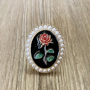 Könnte beinhalten: Ovale Brosche mit schwarzem Hintergrund, auf dem eine rote Rose mit grünen Blättern abgebildet ist. Die Brosche ist von einem goldenen Rahmen und kleinen, weißen Kunstperlen umgeben. Ein klassisches Blumenaccessoire.