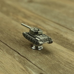 Könnte beinhalten: Silberner Tank-förmiger Schrank-Knopf mit detailliertem Design. Der Knopf ist auf einer runden Basis montiert.