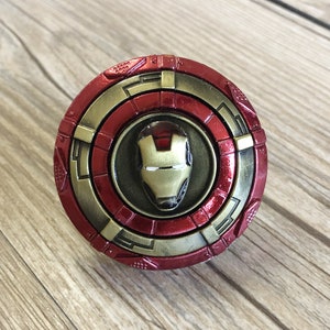 May include: A red and gold metal spinning top with a circular design and a gold Iron Man face in the centre.