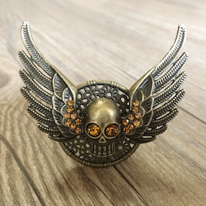 Puede incluir: Un cráneo de metal dorado con ojos de cristal naranja, rodeado de alas de filigrana.