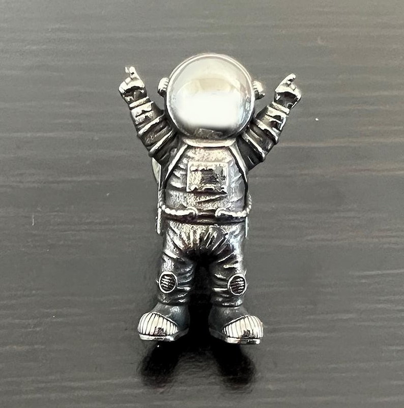 May include: A silver astronaut figurine with raised arms and a clear plastic helmet. The figurine is standing on a dark surface.