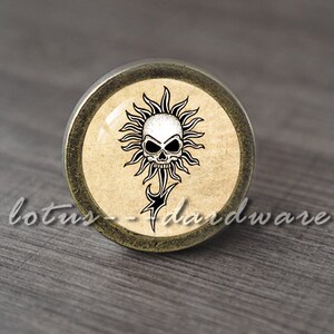 May include: A round, bronze-coloured cabinet knob with a skull and sun design. The skull is white and the sun is black. The background is a light brown, textured paper.