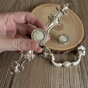 以下が含まれることがあります： 真珠のような中心部を持つ、装飾的なハンドルとノブを備えた銀色のキャビネットハードウェアセット。 セットには2つのハンドルと2つのノブが含まれており、ヴィンテージの美学を示しています。 ハードウェアは木製の表面に表示されています。