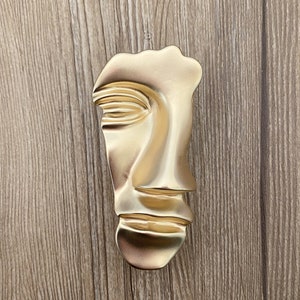 May include: A gold-colored abstract face sculpture with a stylized nose and mouth. The sculpture is made of a smooth, shiny material.