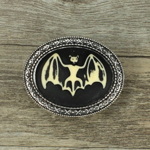 May include: A silver belt buckle with a black and white bat design. The bat is in a flying position with its wings spread out.