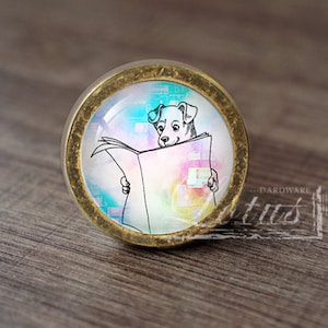 May include: A round, gold-coloured cabinet knob with a glass insert featuring a black and white illustration of a dog reading a newspaper. The background of the illustration is a watercolour-style design with shades of pink, blue, and yellow.