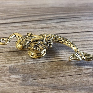 K&ouml;nnte beinhalten: Goldfarbener Meerjungfrau-T&uuml;rknopf mit detailliertem Design. Die Meerjungfrau ist zusammengerollt, ihr Schwanz ragt nach rechts.