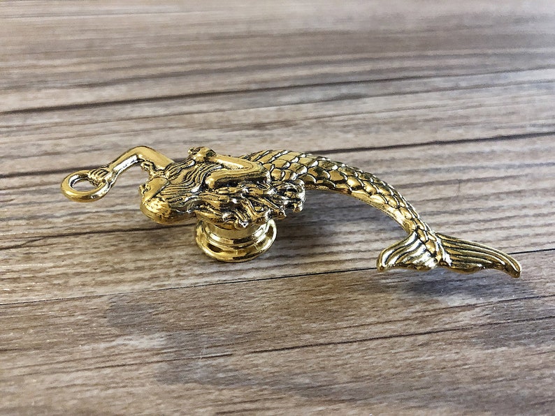 K&ouml;nnte beinhalten: Goldfarbener Meerjungfrau-T&uuml;rknopf mit detailliertem Design. Die Meerjungfrau ist zusammengerollt, ihr Schwanz ragt nach rechts.