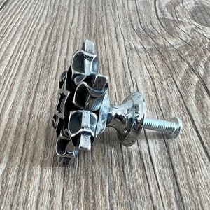 Puede incluir: Un pomo de armario de metal plateado con un dise&ntilde;o geom&eacute;trico &uacute;nico. El pomo tiene una base redonda y un tornillo para fijarlo a una puerta de armario.
