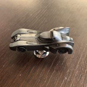 Może przedstawiać: Srebrna metalowa figurka Batmobilu z franczyzy Batman. Figurka jest szczegółowo wykonana z czarnym wykończeniem i ma czarną podstawę.