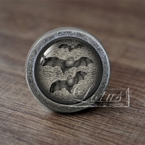 May include: A silver-toned round cabinet knob with a glass insert featuring a black and white image of three bats. The knob has a slightly distressed finish.