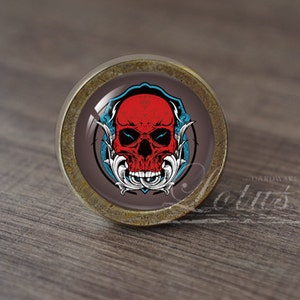 Puede incluir: Un pomo de armario redondo de latón antiguo con un inserto de vidrio que presenta una calavera roja con acentos azules y blancos. La calavera está rodeada por un diseño floral decorativo.