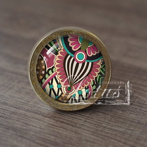 May include: A round, brass-coloured cabinet knob with a colourful, abstract design. The design features pink, blue, green, and black colours.