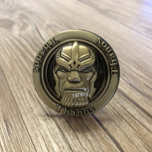 Könnte beinhalten: Eine goldfarbene Metallmünze mit einem erhabenen Bild von Thanos' Gesicht. Die Münze trägt die Inschriften "Soul" und "Thanos".