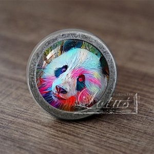 Puede incluir: Pomo de armario redondo de metal con un diseño artístico y colorido de un panda. La cara del panda está representada en tonos azules, rosas y rojos. El pomo está sobre una superficie de madera. Artículo decorativo.