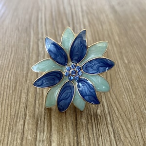 Pode incluir: Uma flor de esmalte azul e azul claro com um centro de strass azuis. A flor é uma peça decorativa.