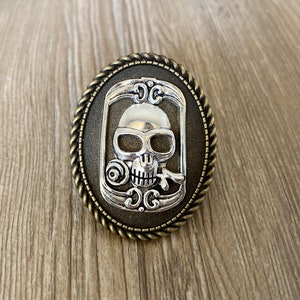 Puede incluir: Hebilla de cinturón ovalada de metal con un diseño de calavera plateada. La calavera está sobre un fondo oscuro dentro de un marco decorativo, rodeada por un borde en forma de cuerda. La hebilla tiene una estética vintage y gótica.