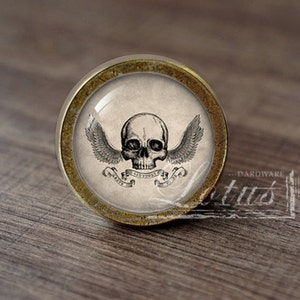 May include: A round, brass-colored cabinet knob with a glass insert featuring a black and white illustration of a skull with wings and a banner that reads "Memento Mori".