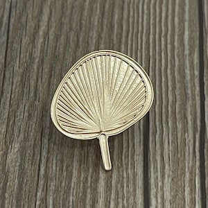 以下が含まれることがあります： 扇形の葉の形をした金色の金属製のピン。ピンは、中心から放射状に伸びる線で詳細なデザインが施されています。