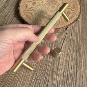 May include: A gold-colored cabinet pull and knob set. The long pull has a textured, diamond-patterned center and two perpendicular handles. The knob has a matching textured surface. The hardware is shown on a wooden surface.