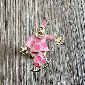 Peut inclure: Une broche de clown rose et or avec des pois blancs. Le clown porte un chapeau et a une expression espiègle.