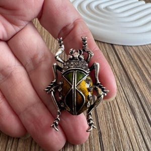 Peut inclure: Broche argentée en forme de scarabée avec un design détaillé. Le corps du scarabée présente un motif vert et jaune, orné de petits cristaux. La broche a des pattes et des antennes complexes.