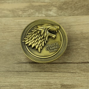 Puede incluir: Un pin de metal dorado con una imagen detallada de una cabeza de lobo huargo y el texto "Winter is Coming Stark".