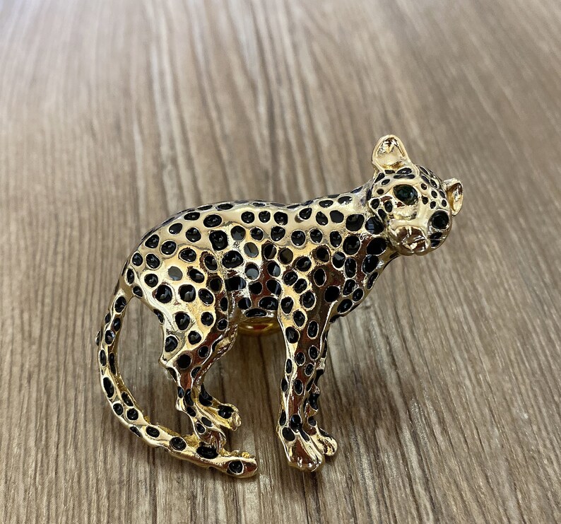 Peut inclure: Une broche en forme de l&eacute;opard dor&eacute;e avec des taches noires. Le l&eacute;opard a des yeux verts et un design d&eacute;taill&eacute;.