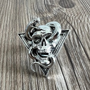 May include: A silver skull with a snake wrapped around it, set in a triangular frame. The skull has a menacing expression with a wide open mouth and sharp teeth.