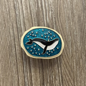 May include: A gold-toned pin with a blue enamel background and white dots. A black whale with a white belly is swimming in the center of the pin.